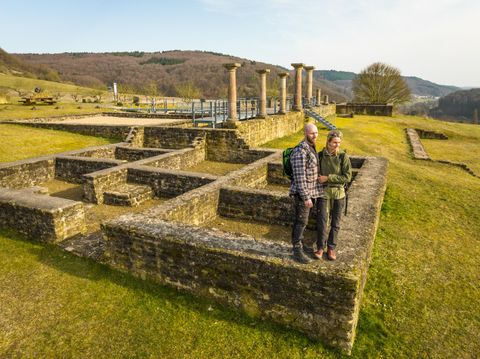 Eine archäologische Stätte mit Ruinen und Säulen im Hintergrund. Zwei Personen stehen in der Nähe der historischen Überreste.