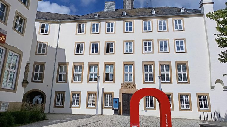 Ein modernes Gebäude mit vielen Fenstern und einem roten Kunstwerk in der Vordergrund. Der Himmel ist blau und teilweise bewölkt.