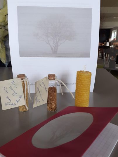A series of small jars with ingredients and a beeswax candle on a table. In the background, there is an image of a tree in a foggy setting.