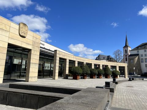 Ein modernes Gebäude mit Glasfassade und Pflanzen vor dem Eingang. Im Hintergrund ist ein Kirchturm und ein klarer blauer Himmel zu sehen.
