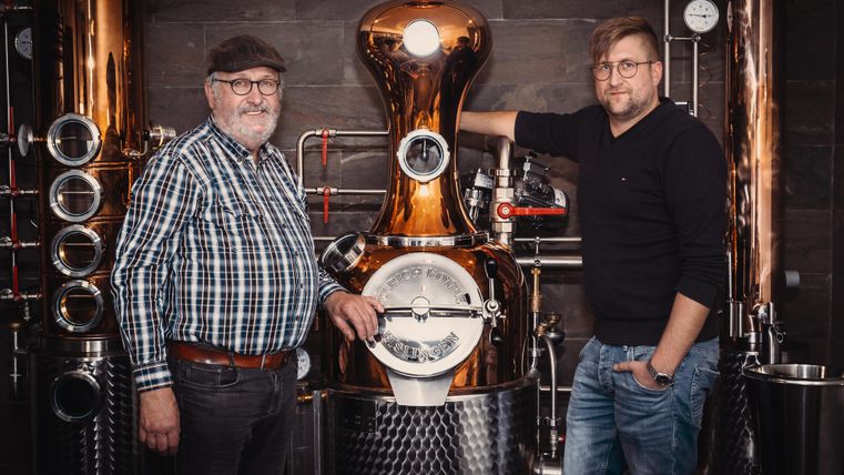 Two men stand next to a stylish distillation plant. They smile at the camera and the plant shines in copper and steel.