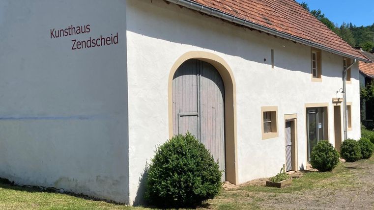 Une maison d'art avec une façade blanche et un toit rouge. L'entrée est décorée d'une grande porte en bois et de petits buissons.