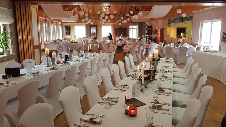 Ein festlich dekorierter Saal mit langen weißen Tischen und eleganten Stühlen. Die Atmosphäre ist einladend mit einer warmen Beleuchtung und dekorativen Elementen.