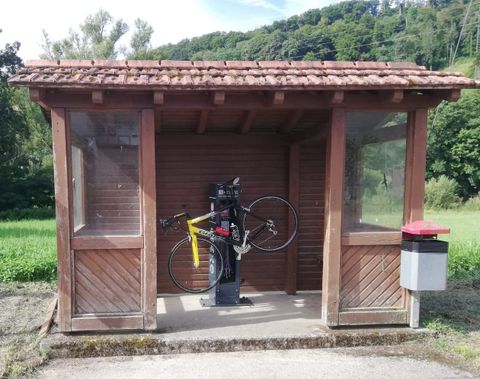 Eine Holzhütte mit einem Radständer und einem gelben Fahrrad. Neben der Hütte steht ein Mülleimer, umgeben von einer grünen Landschaft.