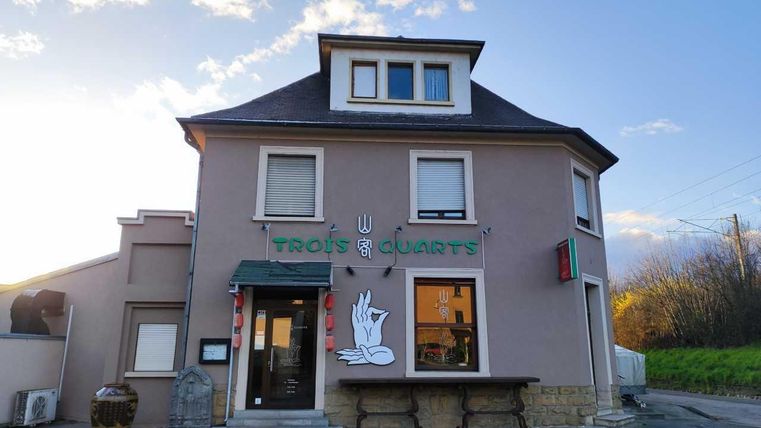 A modern restaurant building with a clear sign showing the name "Trois & Quarts." The facade is simple and inviting, surrounded by a tranquil landscape.