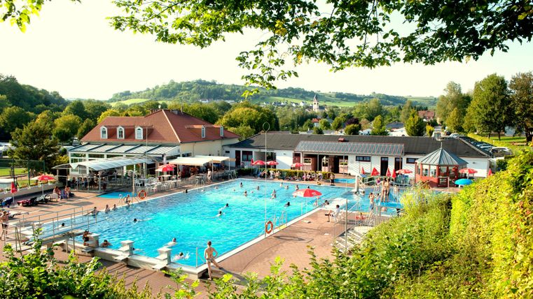 A swimming pool with a large basin and many guests. In the background, green spaces and buildings are visible.