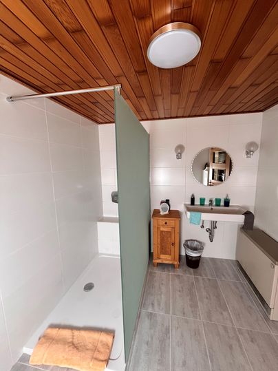 Een moderne badkamer met een lichte douchehoek en een houten plafond. Op de wastafel staan verzorgingsproducten en een kleine houten kast.