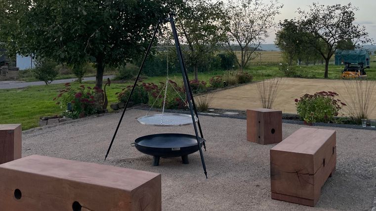 Een gezellige buitenruimte met een kampvuurplek en verschillende houten banken. Omgeven door bomen en een groene weide biedt de plek een uitnodigende sfeer.