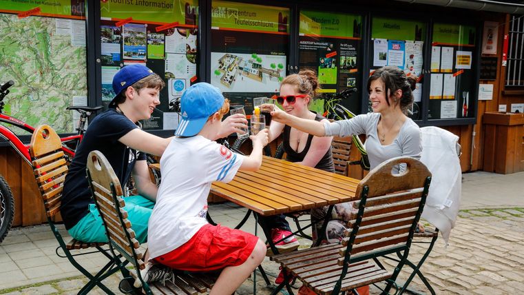 Un groupe de quatre personnes est assis à une table en plein air et trinque avec leurs boissons. À l'arrière-plan, des vélos et des plans d'information sont visibles.