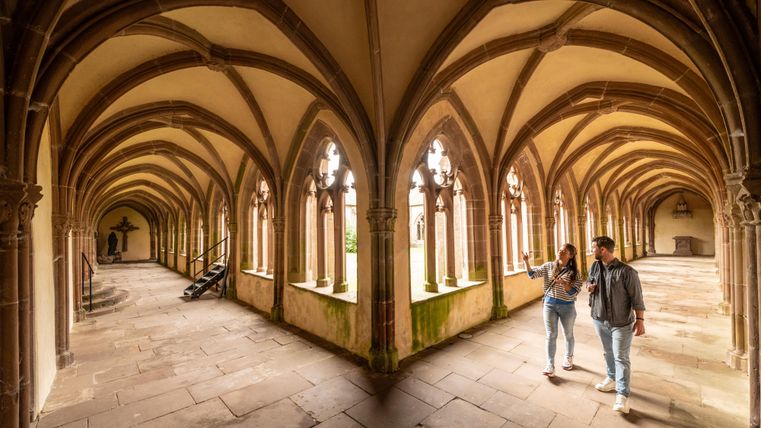 A historic cloister with high arches and strong columns. Two people are strolling leisurely through it.