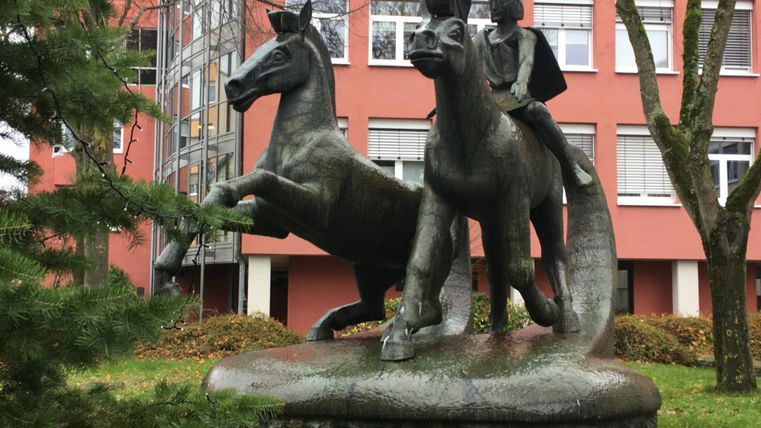 A statue of a rider on two rearing horses. In the background, green trees and a red building are visible.