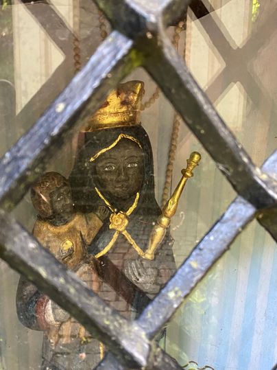 A statue of a protective saint with a child in her arms. The figure wears a crown and holds a scepter, behind a grating window.
