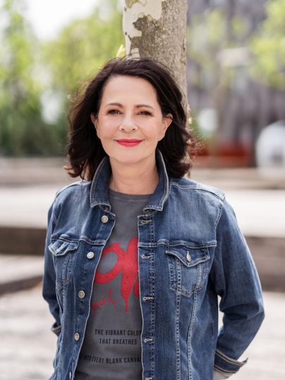 Een vrouw staat glimlachend voor een boom. Ze draagt een spijkerjack en een T-shirt met print.