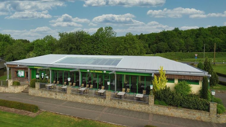 Ein modernes Gebäude mit großen Fenstern und einem hellen Dach. Es liegt inmitten einer grünen Landschaft und hat eine Terrasse mit Sitzplätzen im Freien.