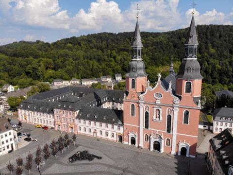 An impressive church with two towers, surrounded by green hills. In the foreground, there is a square with trees and historic buildings.
