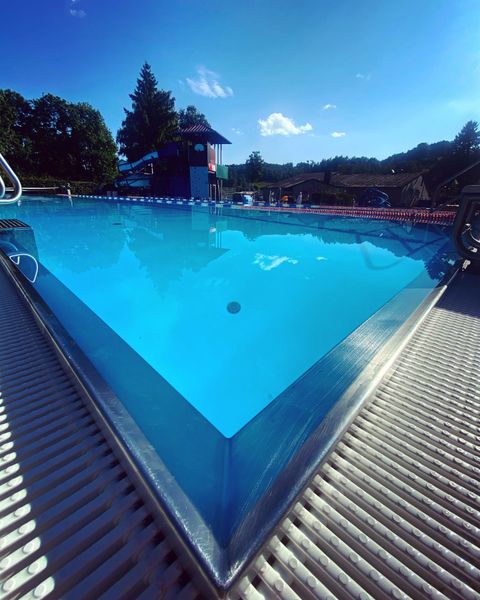 Une piscine claire sous un ciel bleu, entourée d'arbres. En arrière-plan, on aperçoit une aire de jeux.