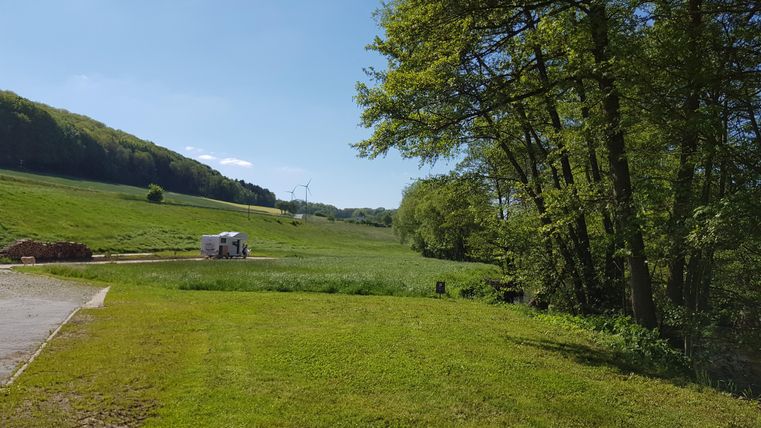 A green meadow with trees and a clear blue sky. In the background, hills and a camper van are visible.