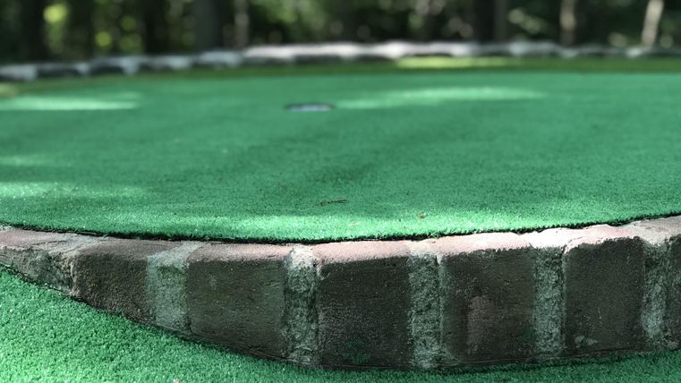 A mini-golf course with green artificial grass and a completely decorated stone border. The background shows a wooded environment.