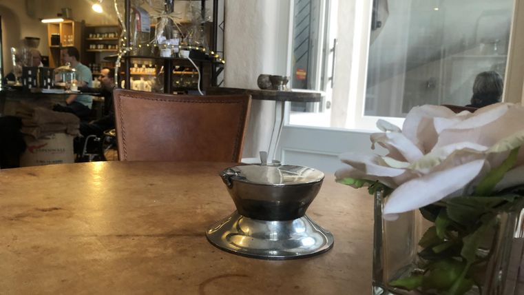 Une table dans un café avec une cafetière en argent et un vase de fleurs. À l'arrière-plan, d'autres clients et une ambiance chaleureuse sont visibles.