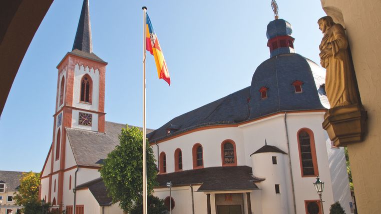 Een schilderachtige kerk met een hoge toren en een kleurrijk dak. Op de voorgrond staat een vlag en er zijn groene bomen te zien.