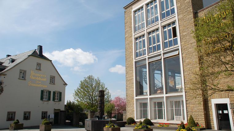 Une place lumineuse avec des bâtiments modernes et traditionnels.
Des arbres et des jardinières fleurissent offrent une atmosphère accueillante.