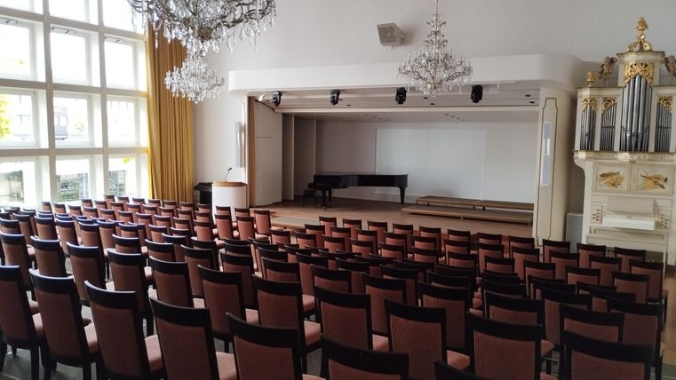 Une grande salle avec des rangées de chaises et un piano sur scène. Les murs sont clairs et il y a des lustres élégants.