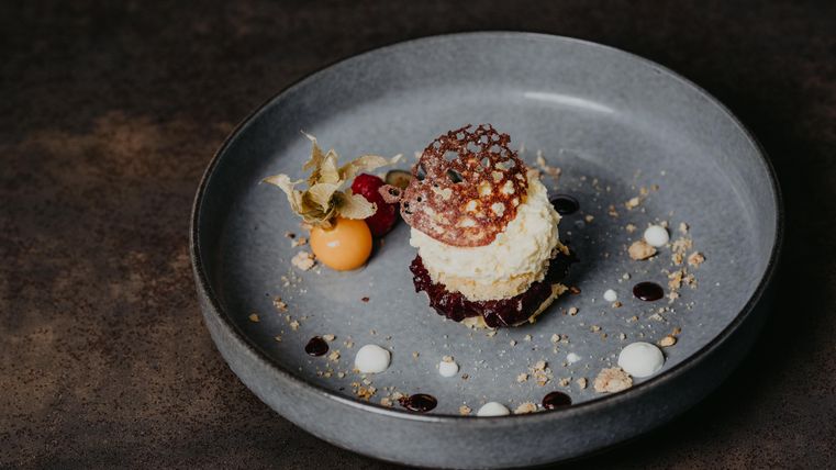 An artistically arranged dessert on a gray plate. It consists of a cream, fruits, and decorative sauces.