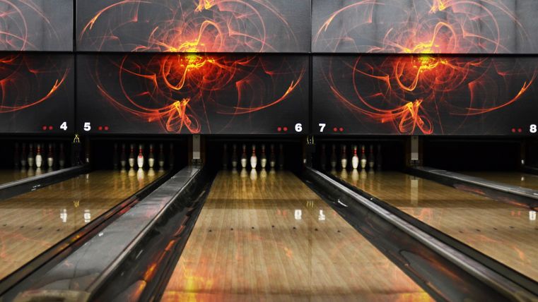Eine Bowlingbahn mit glänzenden Holzoberflächen und elektronischen Anzeigetafeln im Hintergrund. Die Wände sind mit einem modernen, abstrakten Design in warmen Farben dekoriert.