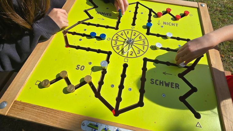A game table with a colorful game board is set up outdoors. Two hands are moving game pieces across the board while the weather looks sunny.