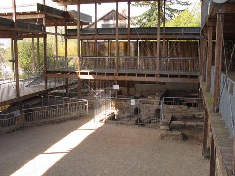 Eine archäologische Ausgrabungsstätte mit Holzstrukturen und einem offenen Bereich. Im Hintergrund sind einige Überreste sichtbar, umgeben von Zäunen.