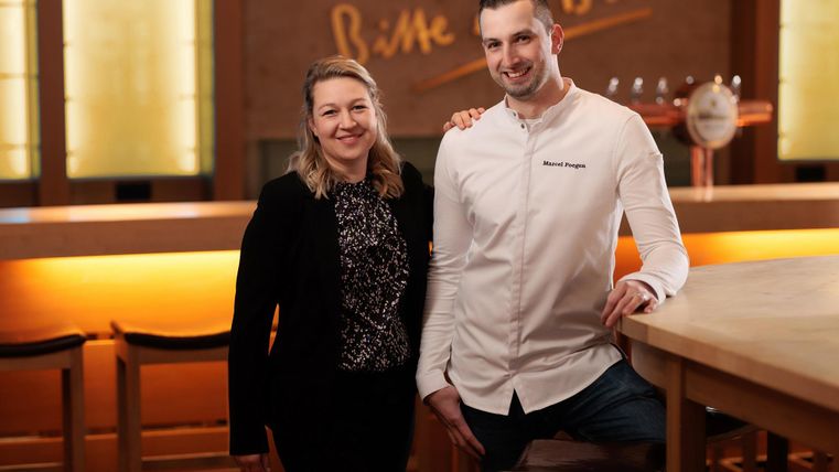 A couple stands smiling at a bar in a restaurant. In the background, a warm light glows.