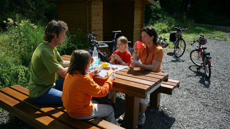 Familie macht Rast an einem Holztisch mit Fahrrädern im Hintergrund.