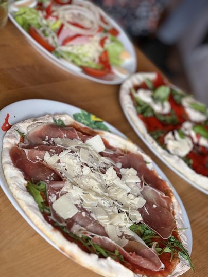 Une délicieuse pizza avec du jambon et de la roquette est posée sur une table. En arrière-plan, on peut voir des salades fraîches.