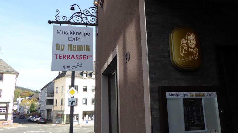 A sign for a music pub and café named "by Namik" is attached to the wall.
It is a quiet street with several buildings in the background.