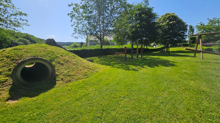 Un grand terrain de jeu vert avec une structure en forme de colline et un tunnel. À l'arrière-plan, il y a des arbres et des équipements de jeu.