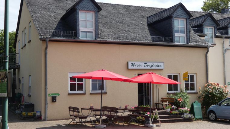 Een gezellig gebouw met een bord dat "Huis Drievuldigheid" toont. Voor het huis staan tafels met rode zonneschermen en bloemen.