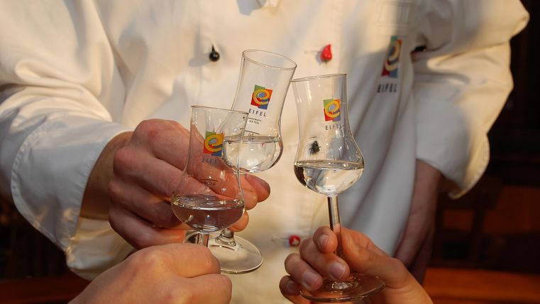 Four glasses with a clear drink are being raised for a toast. In the background, a person in chef's clothing can be seen.