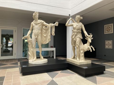 Two large statues stand in a modern room. On the left a man with a cloak, on the right a woman with a deer.