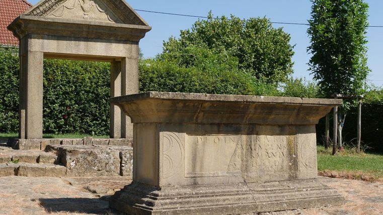Ein historischer Steinaltar steht in einem Garten mit Bäumen im Hintergrund. Über dem Altar befindet sich ein dekorativer Bogen.