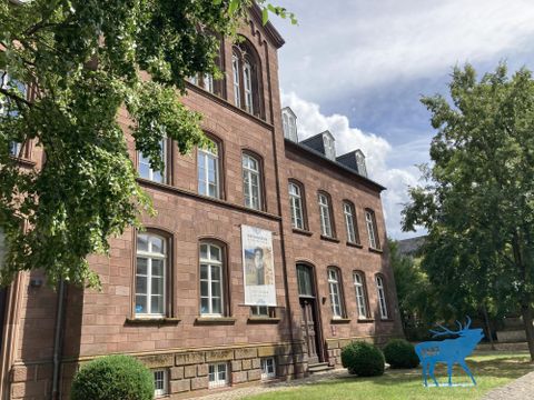 A historical building made of red stone with large windows. Surrounded by trees and green grass under a blue sky.