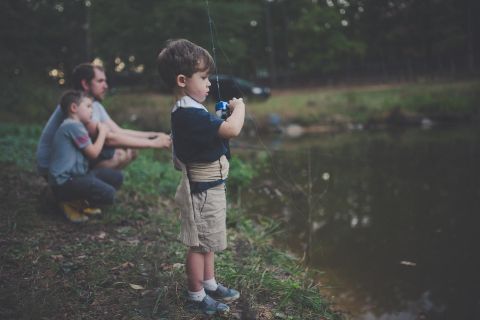 A little boy is fishing at the edge of a body of water. In the background, two adults can be seen fishing as well.