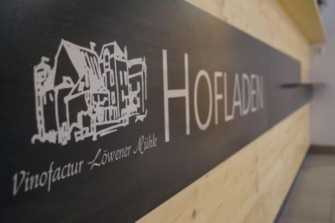 Ein Schild mit der Aufschrift "Hofladen" und einer skizzenhaften Darstellung von Häusern. Das Logo gehört zur Vinofaktur Löwenherz.