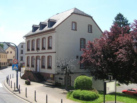 Un bâtiment lumineux à deux étages avec des murs blancs et un toit en pente. Au premier plan, il y a une pelouse verte et quelques arbres.