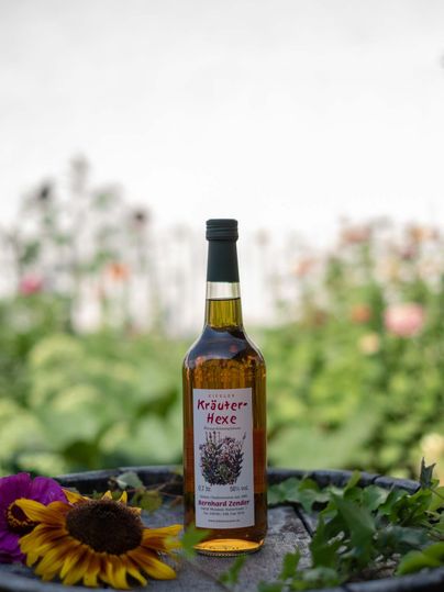 Eine Flasche Kräuterhexe steht auf einem Fass, umgeben von bunten Blumen und Pflanzen. Der Hintergrund zeigt eine grüne Gartenlandschaft.