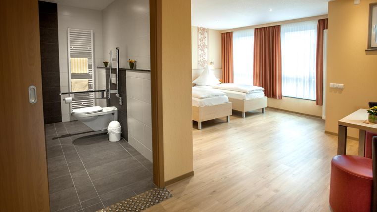 A modern hotel room with two single beds and large windows. In the background, a bathroom with modern sanitary facilities is visible.