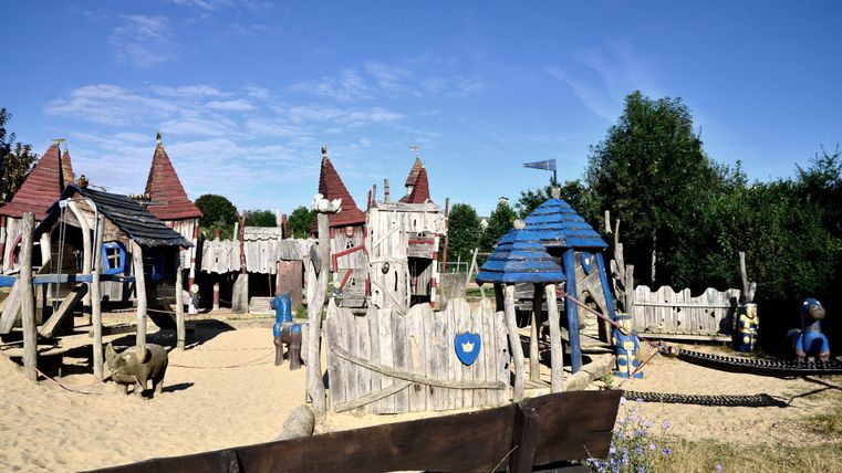 Een speeltuin met een houten speelburg en verschillende glijbanen. Omgeven door groene bomen en een heldere lucht.