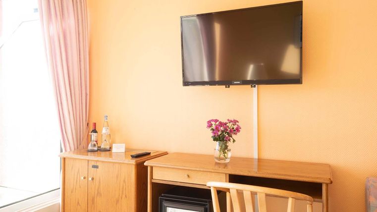 Ein gemütlicher Raum mit einem Fernseher an der Wand und einem Holztisch. Auf dem Tisch steht eine Vase mit Blumen und daneben ein Kühlschrank.