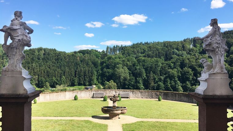 Eine wunderschöne Gartenlandschaft mit einem Brunnen in der Mitte und Skulpturen an den Seiten. Im Hintergrund sind grüne Hügel und ein blauer Himmel zu sehen.
