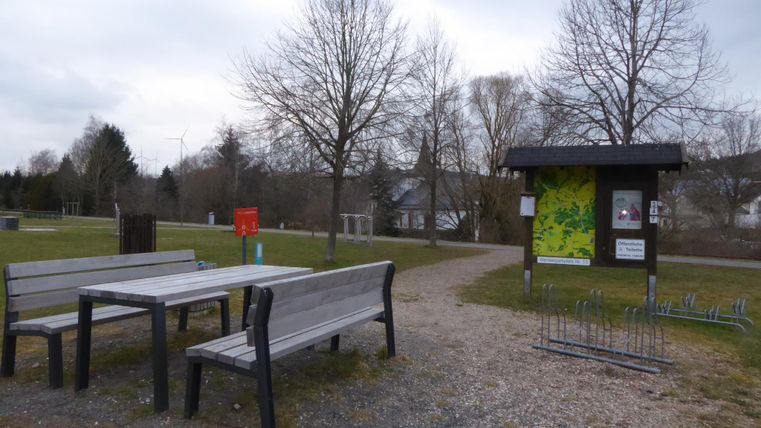 A quiet park with wooden tables and benches. In the background, there is an information kiosk and some trees.