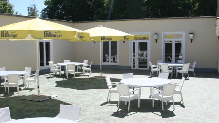 A bright terrace with tables and chairs. Umbrellas provide shade and the surroundings are green.